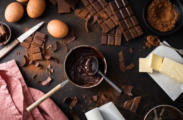 A pot of melted chocolate on a surface surrounded by pieces of chocolate, cocoa powder, and other baking essentials.