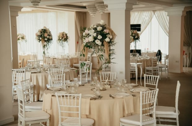 An event venue with elegant tables, chairs, napkins, and dinnerware. Each table has tall floral centerpieces.