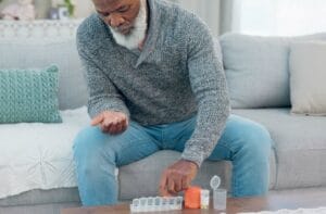 An older African American man organizing his medication in a pill organizer. He’s at home and sitting on a couch.