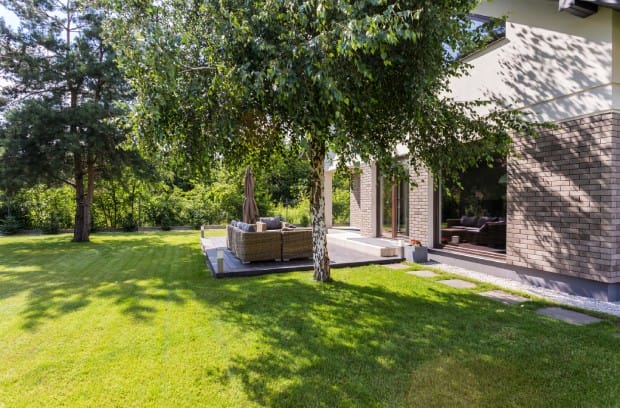 A backyard contains a small concrete patio with an outdoor couch, green grass, concrete pavers, and large trees.