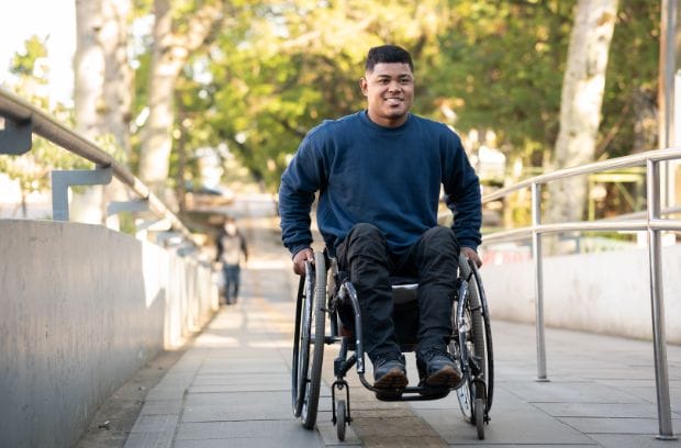 A wheelchair user in a blue sweater and black jeans, rolling up a ramp outside in front of some trees. It's sunny outside.