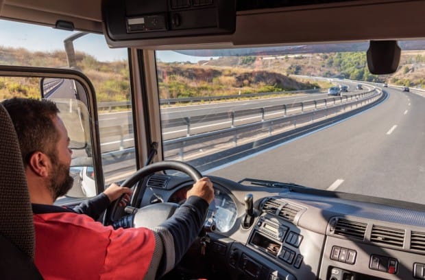 A truck driver sitting in their truck's cab. They drive down a highway, surrounded by desert sand, bushes, and mountains.