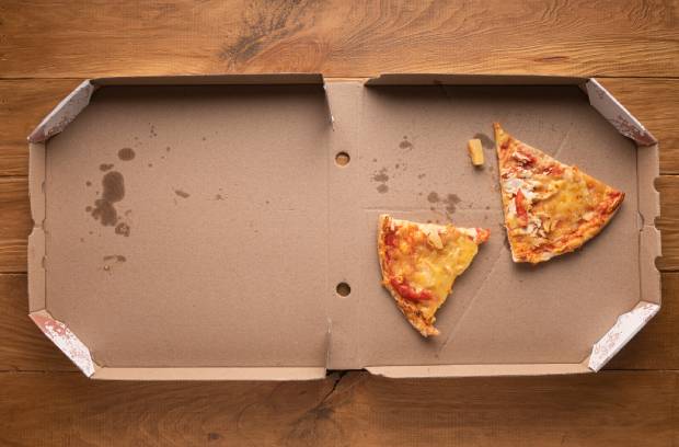 An open pizza box sits on a wooden table. It contains two slices of pizza, a pineapple bit, and grease stains.
