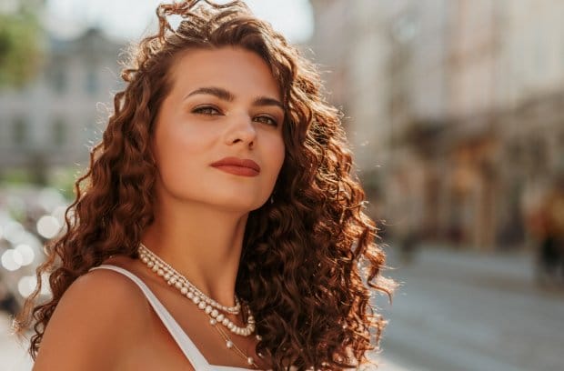 A young woman with curly auburn hair wears a white shirt. She is wearing different lengthed pearl necklaces.