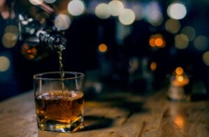 A hand is pouring whiskey from a glass bottle into a small, square glass. The glass sits on a wooden table.