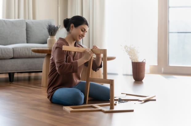 A woman sits on a living floor and builds a wooden chair. She smiles and focuses on joining two wood pieces.