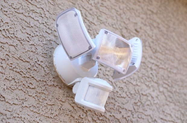 A white motion sensor light contains three separate lights. It sits on a tan wall with various grooves.