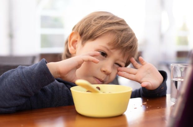 A little boy sitting at the dinner table with a small yellow bowl and spoon in front of him. He's rubbing his eyes.