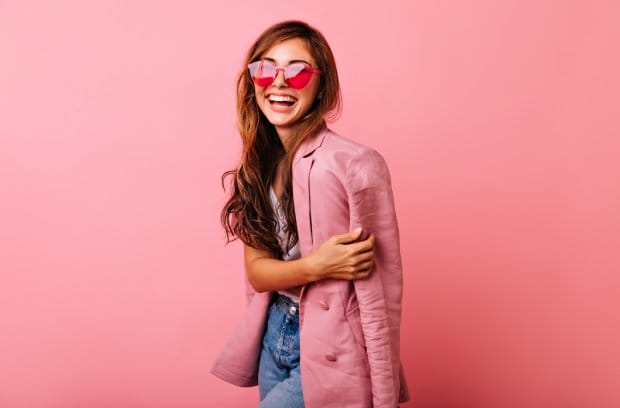 A young woman is wearing a pink blazer, jeans, and pink sunglasses. She's smiling and standing against a pink background.