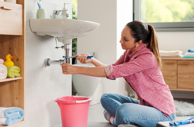 A woman sits on the floor using a blue wrench to tighten an exposed metal pipe under a white sink in the bathroom.