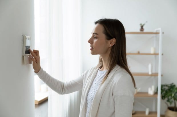 A female homeowner standing at her home's wall-mounted thermostat. She's adjusting the temperature settings.
