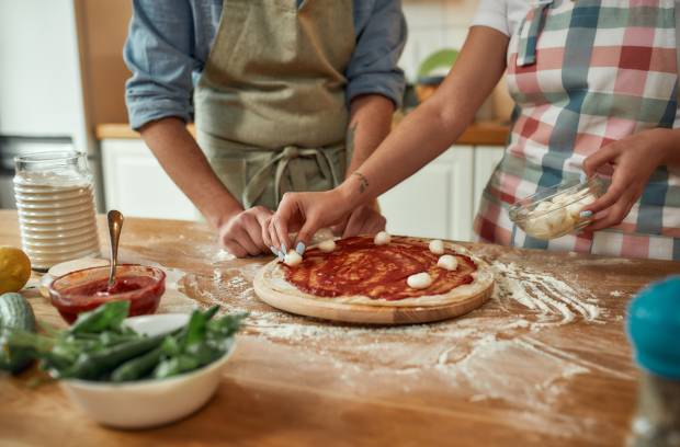 A couple wearing aprons placing mozzarella balls on a homemade pizza in their kitchen surrounded by other ingredients.