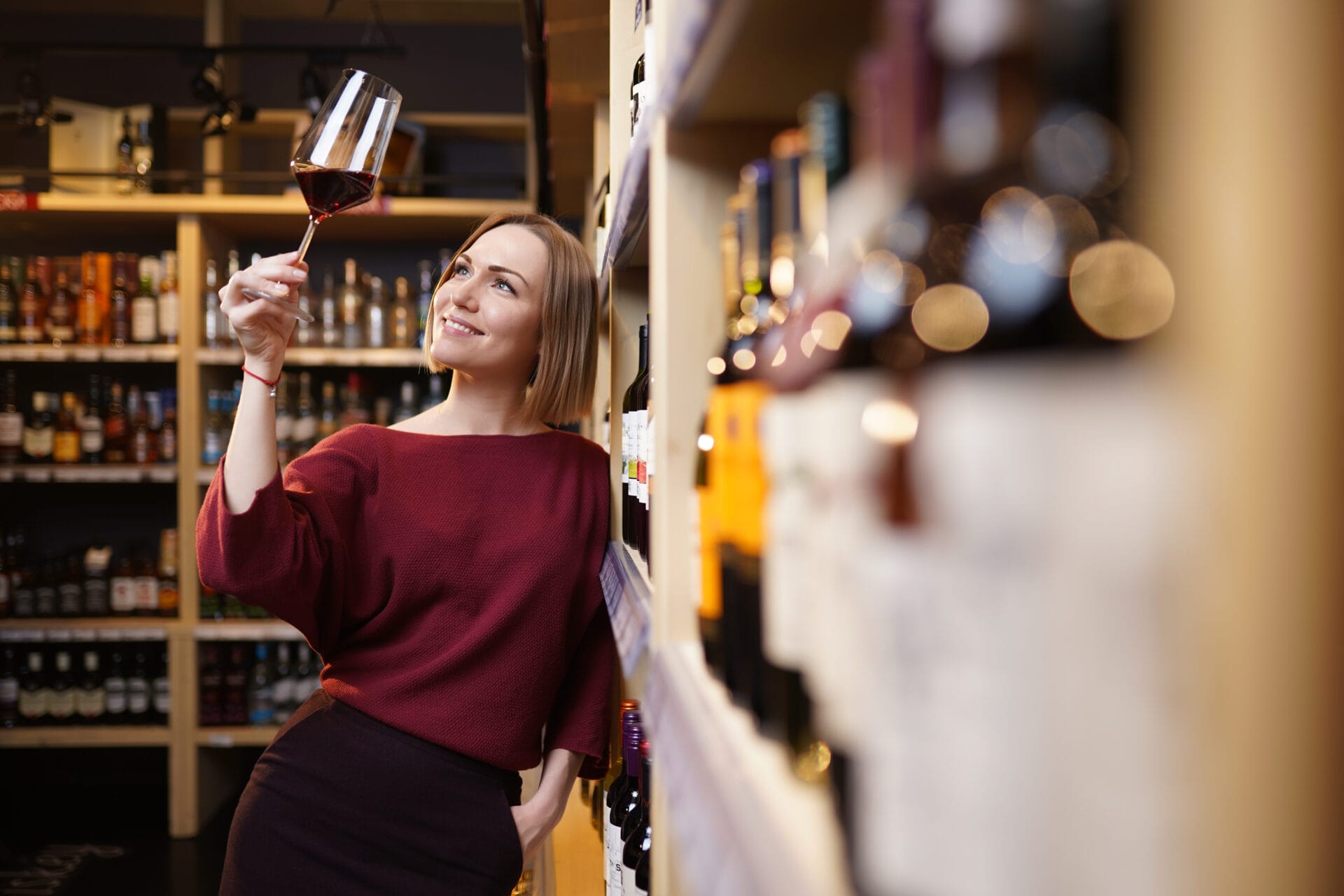 A woman wearing a red sweater holds a glass of red wine up to the light, inspecting it next to shelves of wine bottles.