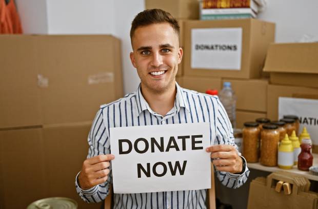 A smiling man holds a sign that says, "Donate Now." Donation boxes and food sit on tables behind him.