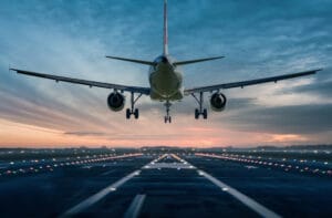 A large, white airplane is landing on an airport runway with lights and white lines. The sun is setting.