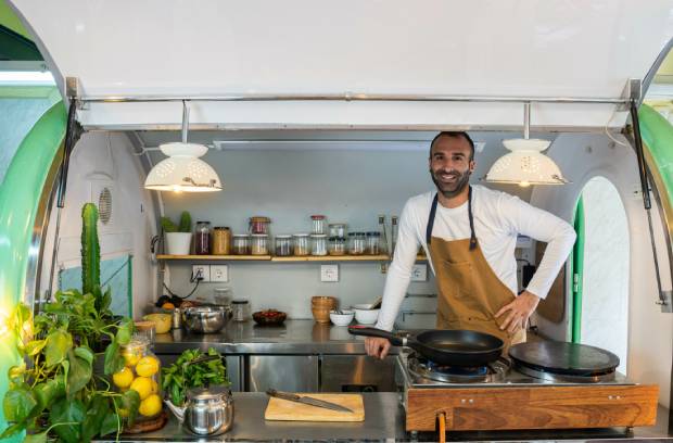 A man smiles while standing inside a food truck. He’s in front of a stove with a pan, and spices are on the shelves behind him.