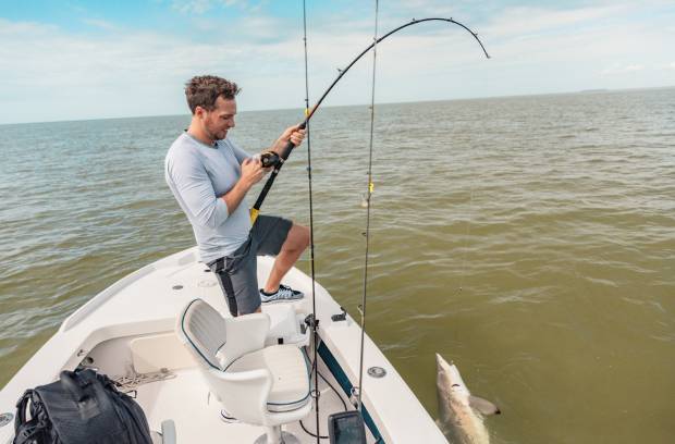 A man stands on the front of a fishing boat holding a fishing pole that has a large fish hooked on the end.