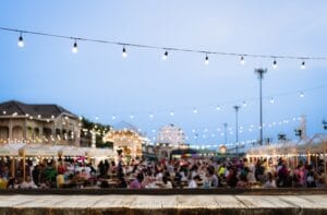 Outdoor string lights hang above a community event at dusk. Guests sit at tables and walk past booths.
