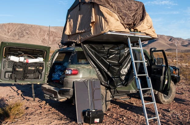 A tan rooftop tent sits on top of a forest-green SUV with the back door open. The vehicle sits in the desert.