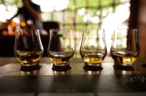 A wooden plank contains four bourbon glasses slightly filled with an amber-colored liquid. The background is blurry.