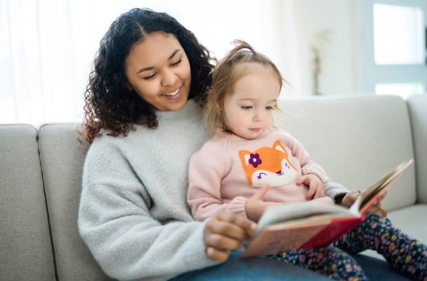 A young woman in a gray sweater reads a picture book to a toddler in a pink sweater sitting on her lap.