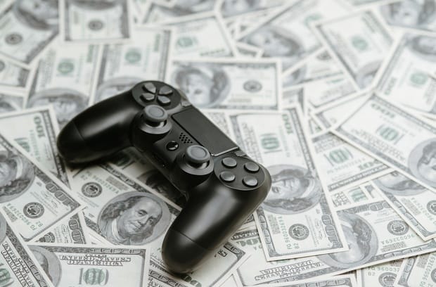 A black modern video game controller sitting on top of a spread out pile of crisp 100 dollar bills.