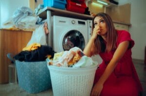 A woman sits next to a laundry basket with clean clothes and holds her nose. A washing machine is behind her.