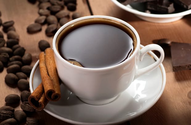 A cup of coffee in a white mug sitting on a white saucer. The coffee is black and the mug is surrounded by loose beans.