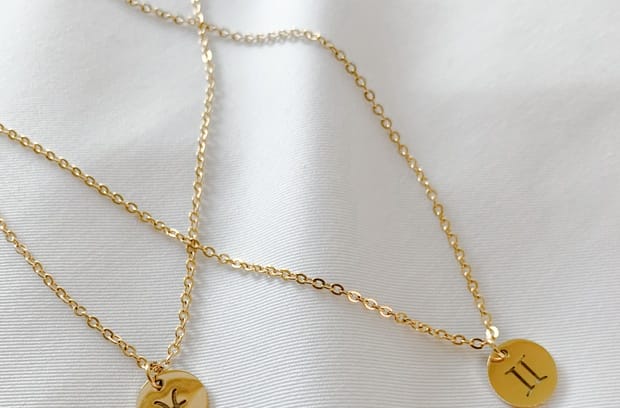 Two gold chain necklaces with zodiac symbols on flat pendants lie next to each other on a piece of white fabric.