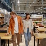 A couple walking down an aisle at the furniture store as they both look at dining room tables and chairs.