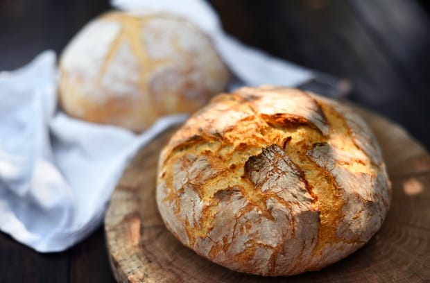 A fresh loaf of sourdough sitting on a wooden cutting board with texture on the crust. There's a second loaf behind it.