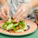 A chef wearing a white apron and clear food prep gloves as they prepare a plate of salad and vegetables.