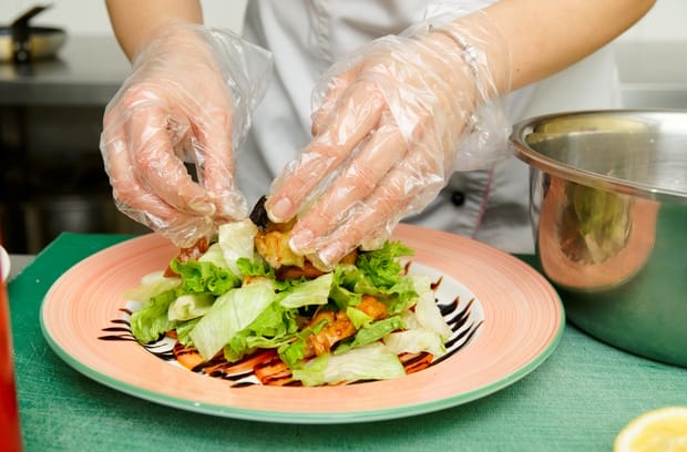 A chef wearing a white apron and clear food prep gloves as they prepare a plate of salad and vegetables.