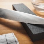 A close-up view shows a chef's knife sitting on a black block next to a small bowl of water on a wooden surface.