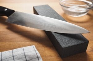 A close-up view shows a chef's knife sitting on a black block next to a small bowl of water on a wooden surface.