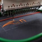 An embroidery machine uses orange thread to create letters on a piece of black fabric in a green embroidery hoop.