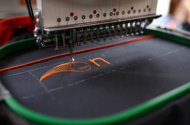 An embroidery machine uses orange thread to create letters on a piece of black fabric in a green embroidery hoop.