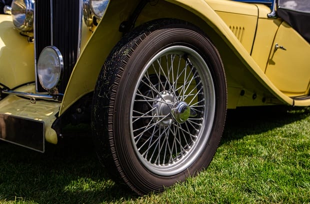 A close-up showcasing the shiny silver spoke wire wheels on a vintage yellow car parked on a patch of grass.