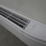 A close-up view shows a white baseboard heater attached to a white wall. The carpet under the heater is gray.