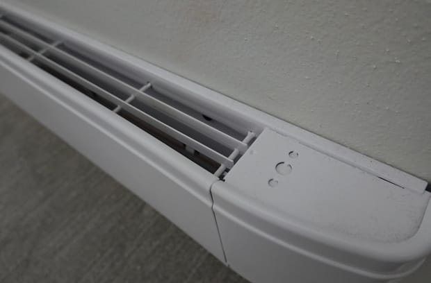 A close-up view shows a white baseboard heater attached to a white wall. The carpet under the heater is gray.