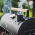 A close-up view shows a black meat smoker with a wooden handle. Smoke is coming out the smoker and the background is blurry.