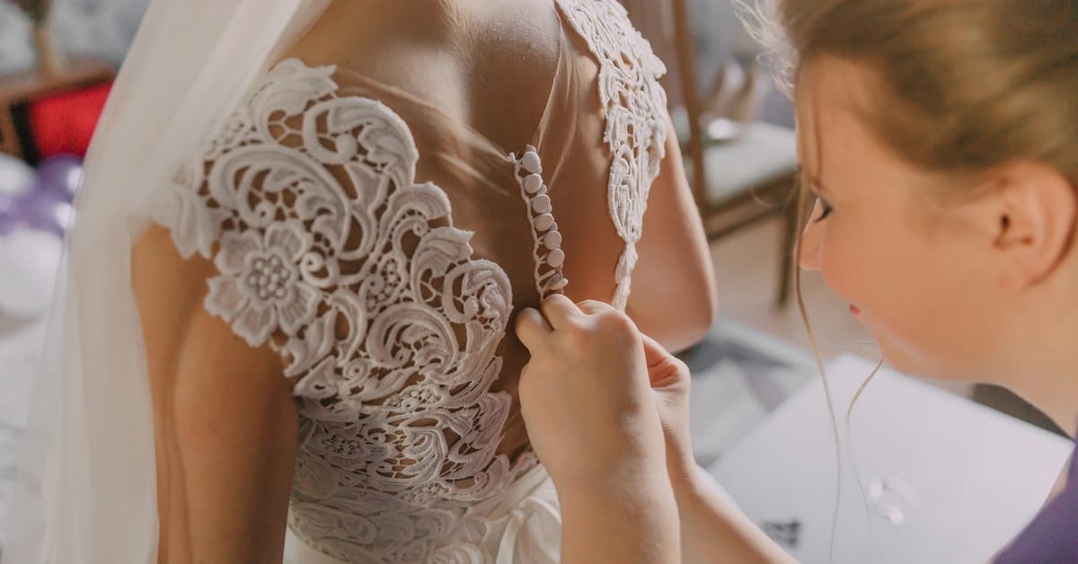 A woman fastens the buttons on the back of a bride's dress. Warm sunlight pours into the room, illuminating them.