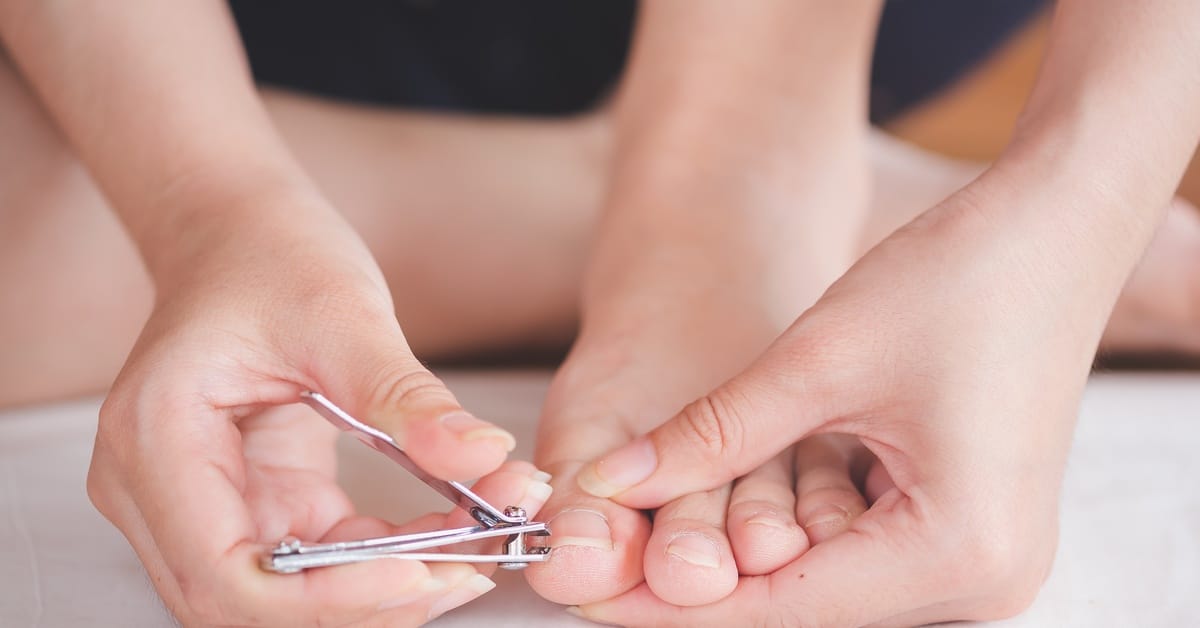 A close-up view shows a person sitting down and using metal nail clippers to trim the nail on their big toe.