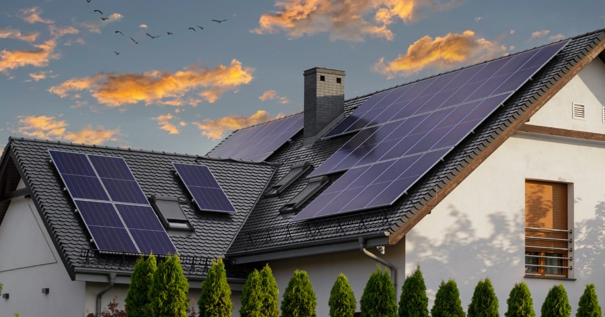 A two-story white home has several solar panels on the roof. The sunset is turning the clouds orange behind the home.