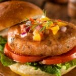 A salmon burger sits on top of two slices of tomato, a leaf of lettuce, and the bottom bun. The top bun sits to the side.