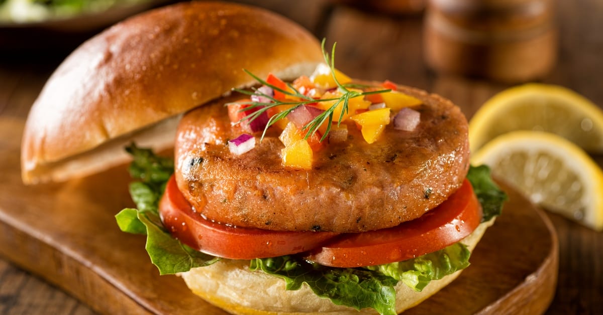 A salmon burger sits on top of two slices of tomato, a leaf of lettuce, and the bottom bun. The top bun sits to the side.