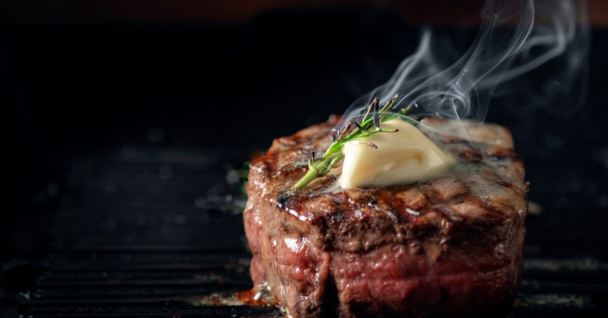 A beef tenderloin rests on a grill with smoke rising from the meat. Butter and a small sprig of rosemary are on top of the beef.