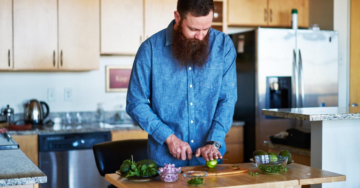 A man is slicing a lime on a small, wooden island in a kitchen. Glass bowls with other ingredients sit on the island.