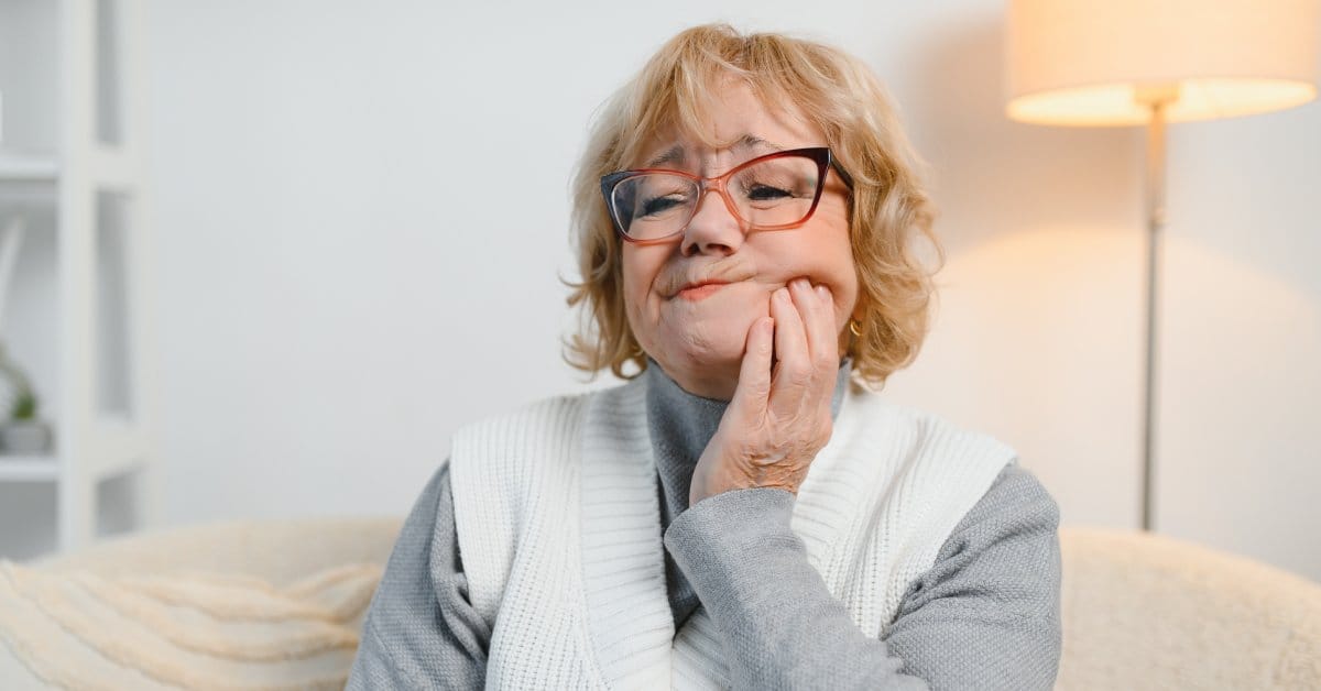 A woman wearing glasses, a gray shirt, and a white sweater vest holds her cheek. A look of pain is on her face.