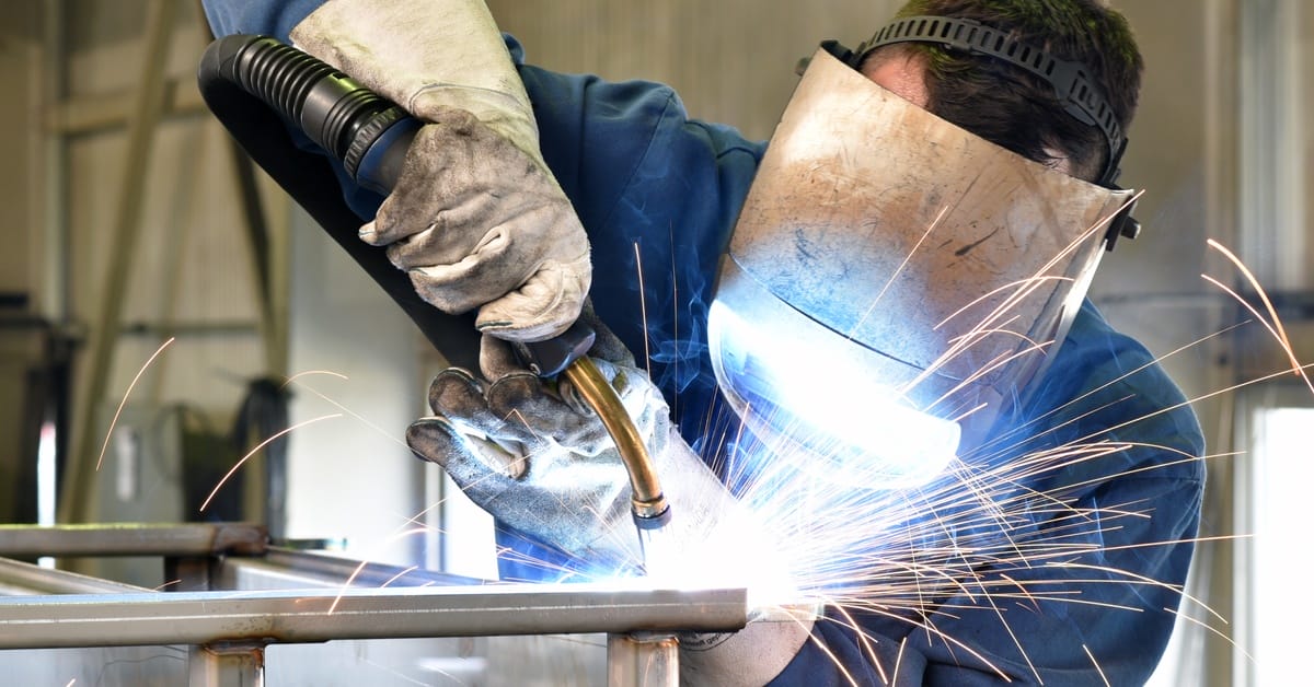 A man wearing a welding mask using a welding tool to work on a steel rod, with sparks flying around him.